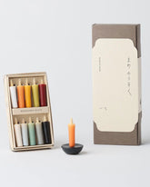 A product shot of a Daiyo mini rice candle set showing a neatly boxed set of 10 earth coloured candles, a small orange candle on a small black circular stand and a tall brown cardboard box with a cream coloured paper slip label. Set on a light grey background. 