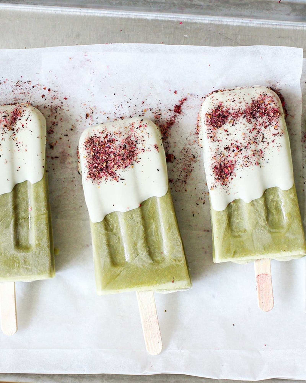 Pink-coloured, Cardamom and Rose spice dust sprinkled across three green and white popsicles on wooden sticks, resting on parchment paper inside a metal baking pan.