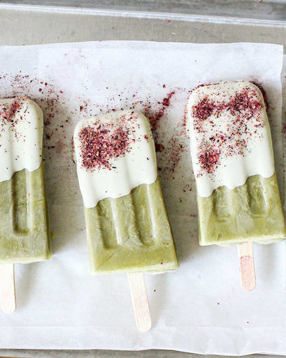 Pink-coloured, Cardamom and Rose spice dust sprinkled across three green and white popsicles on wooden sticks, resting on parchment paper inside a metal baking pan.