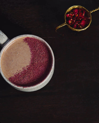 A ceramic mug filled with a latte beverage, the top of the latte is dusted with Cardamom and Rose pink-coloured spice dust in the shape of a moon crescent.