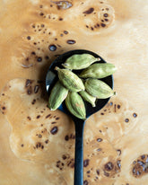 Green cardamom pods resting in a black spoon, set against a textured, wood surface.