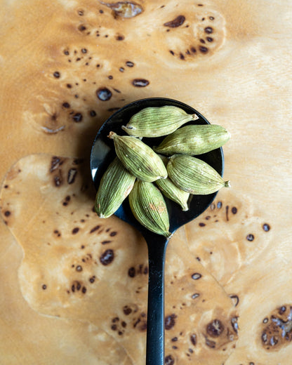 Green cardamom pods resting in a black spoon, set against a textured, wood surface.