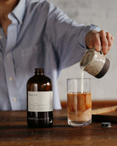 Person is standing at a dark wooden counter pouring milk from a ceramic milk jug into a tall glass with Dona&