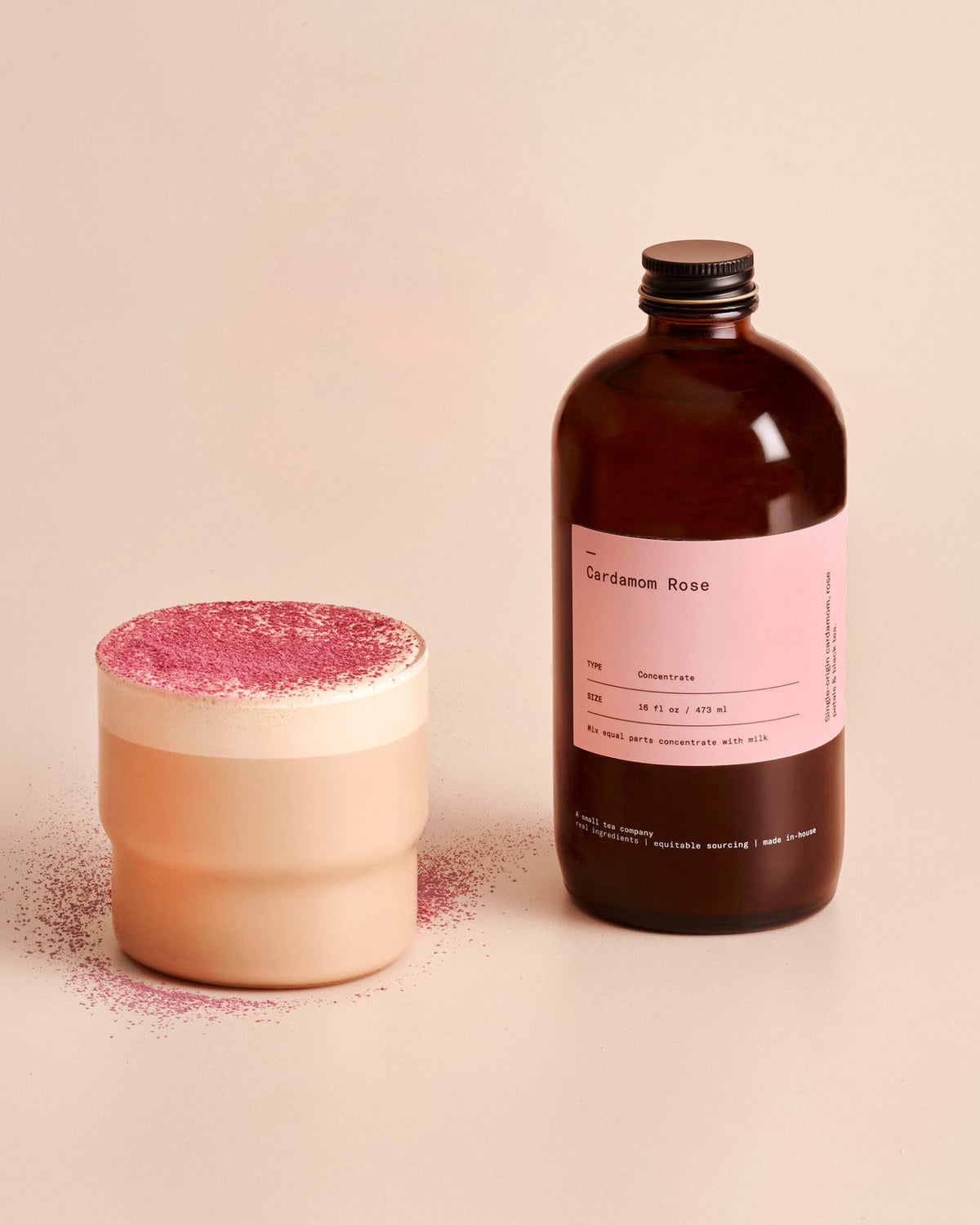 A creamy latte is featured in a glass drinking cup, the latte is dusted with a pink powder. An amber glass bottle labelled Cardamom Rose sits next to the glass. Both sit against a solid cream-coloured background. 