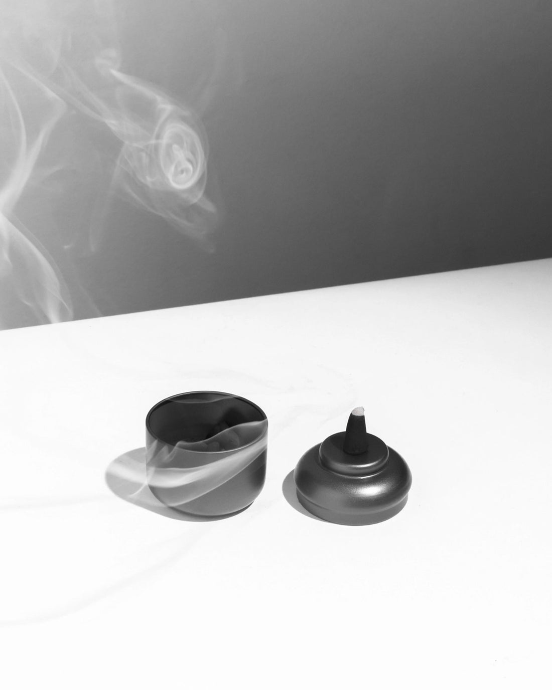 On a white table, a grey-coloured tin canister sits with its lid to the side. The open canister shows contents of black charcoal incense cones, the lid sits next to the open canister, a lit incense cone sits on top of the lid, there is a line of smoke drifting from the tip of the incense cone. 