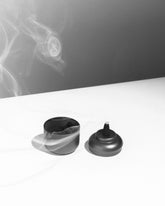 On a white table, a grey-coloured tin canister sits with its lid to the side. The open canister shows contents of black charcoal incense cones, the lid sits next to the open canister, a lit incense cone sits on top of the lid, there is a line of smoke drifting from the tip of the incense cone. 