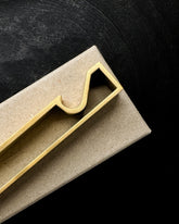 A minimalist, modern geometric bottle opener with a gold brushed metal finish laying flat on a light brown cardboard box on a dark black wood background. 