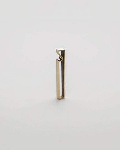 A minimalist, modern geometric bottle opener with a gold brushed metal finish, standing upright on a seamless light grey background.