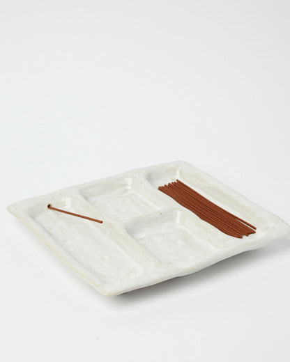 Product shot of a white ceramic platter designed with 4 recessed compartments, one compartment shows a hole in one end with an incense stick resting through the hole. A bundle of incense sticks sits in an another compartment, the other 2 compartments are empty. 