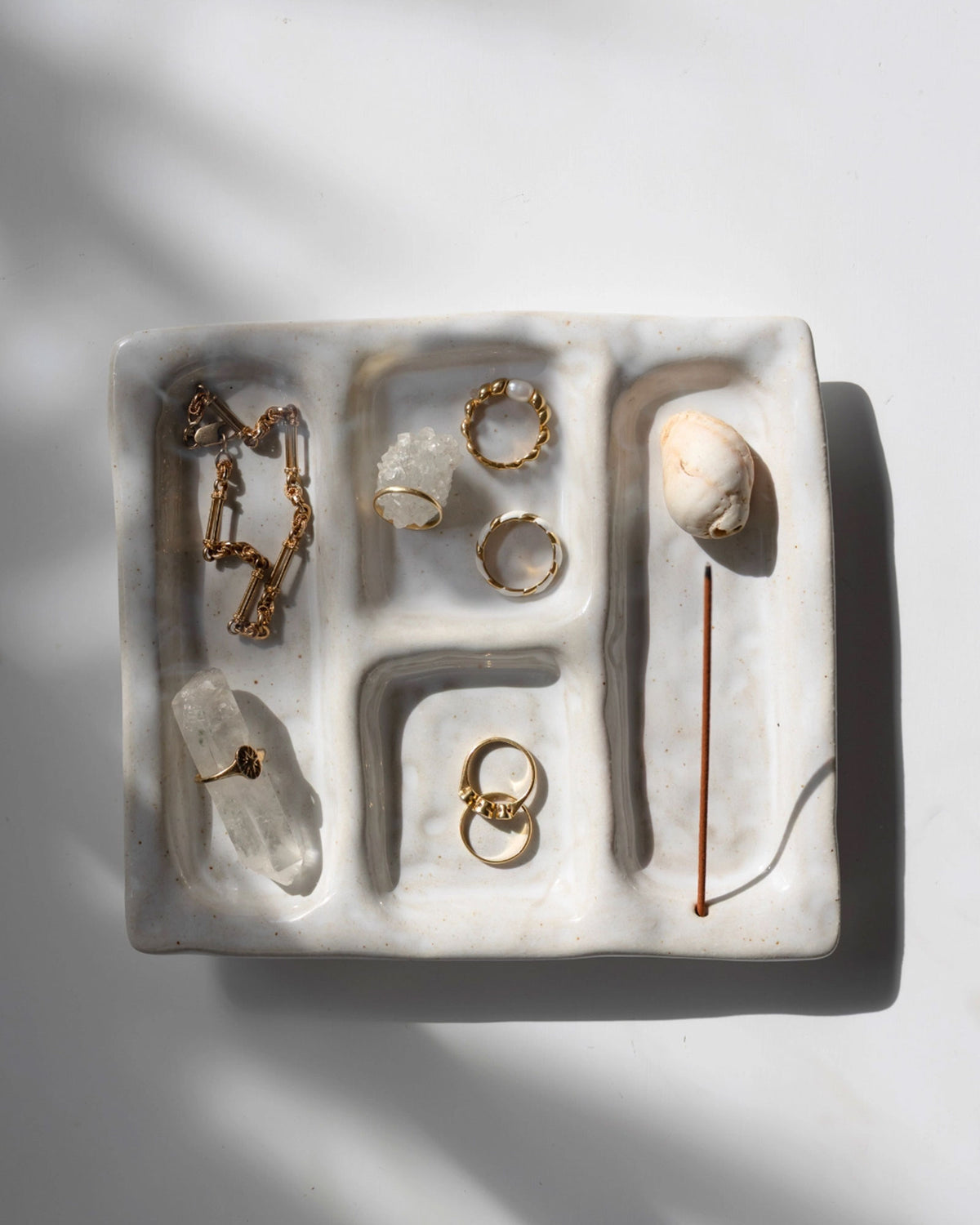 A white ceramic platter with 4 recessed compartments styled with crystals and gold jewellery scattered amongst the compartments. One compartment has a hole inserted at one end to hold an incense stick and collect the falling ash. 