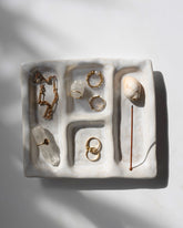 A white ceramic platter with 4 recessed compartments styled with crystals and gold jewellery scattered amongst the compartments. One compartment has a hole inserted at one end to hold an incense stick and collect the falling ash. 