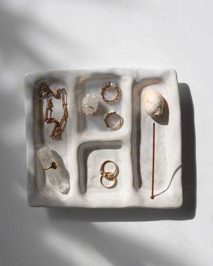 A white ceramic platter with 4 recessed compartments styled with crystals and gold jewellery scattered amongst the compartments. One compartment has a hole inserted at one end to hold an incense stick and collect the falling ash. 