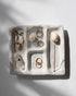 A white ceramic platter with 4 recessed compartments styled with crystals and gold jewellery scattered amongst the compartments. One compartment has a hole inserted at one end to hold an incense stick and collect the falling ash. 