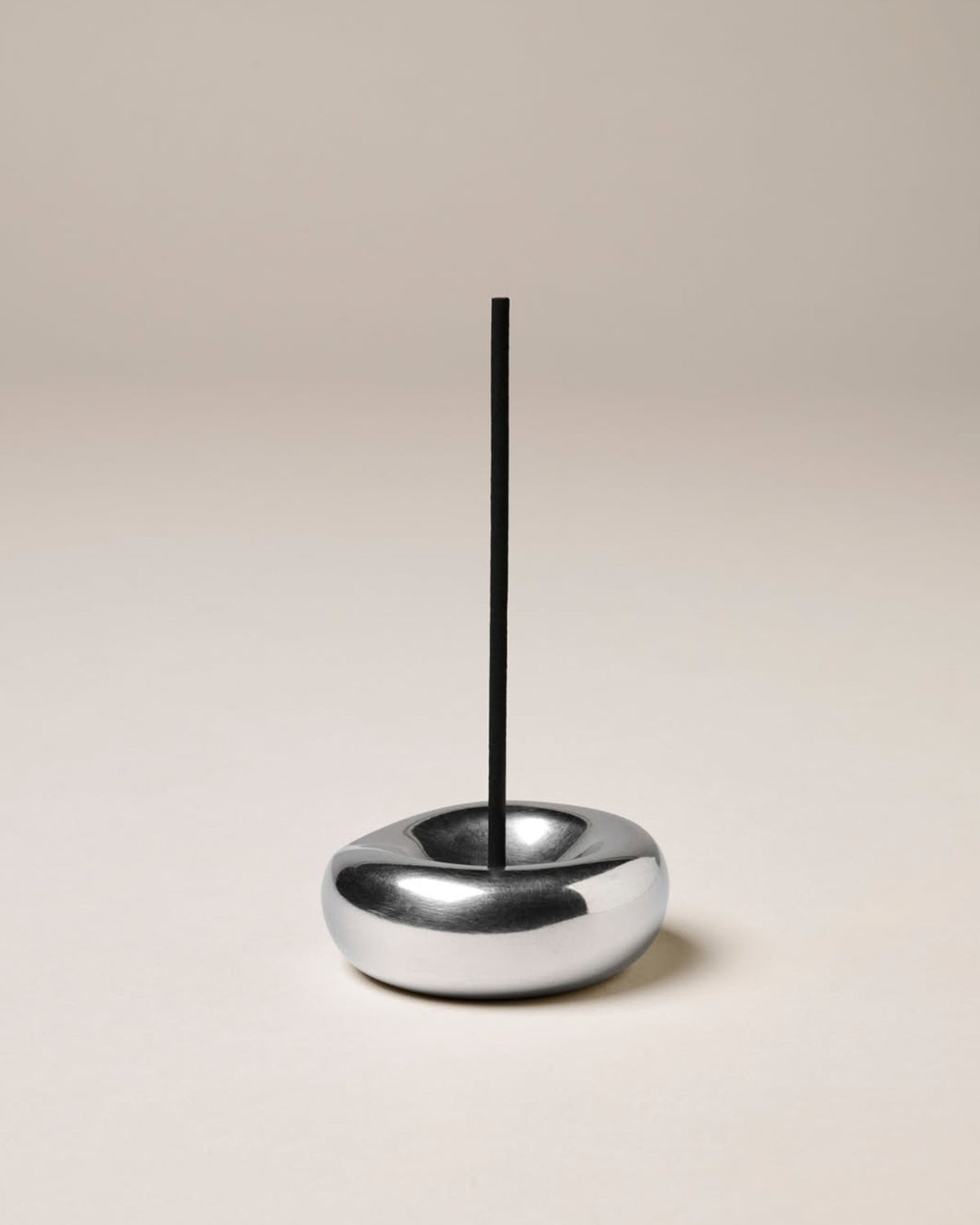 A closeup product shot of a round, sculptural chrome incense holder with a vertical black incense stick placed in the centre of the holder.