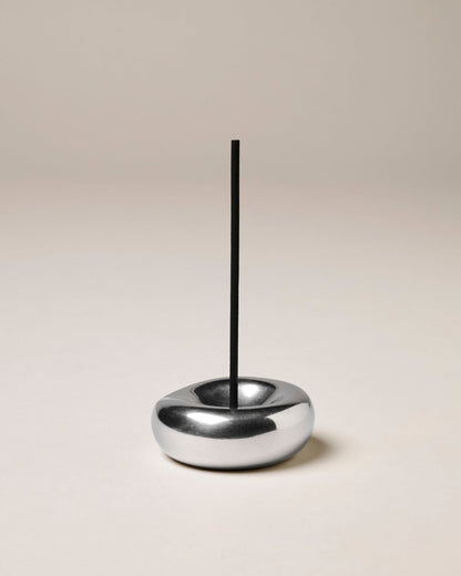 A closeup product shot of a round, sculptural chrome incense holder with a vertical black incense stick placed in the centre of the holder.