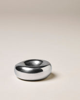 A minimalist, organic-shaped chrome incense holder with a smooth, polished finish, set against a solid warm beige background.