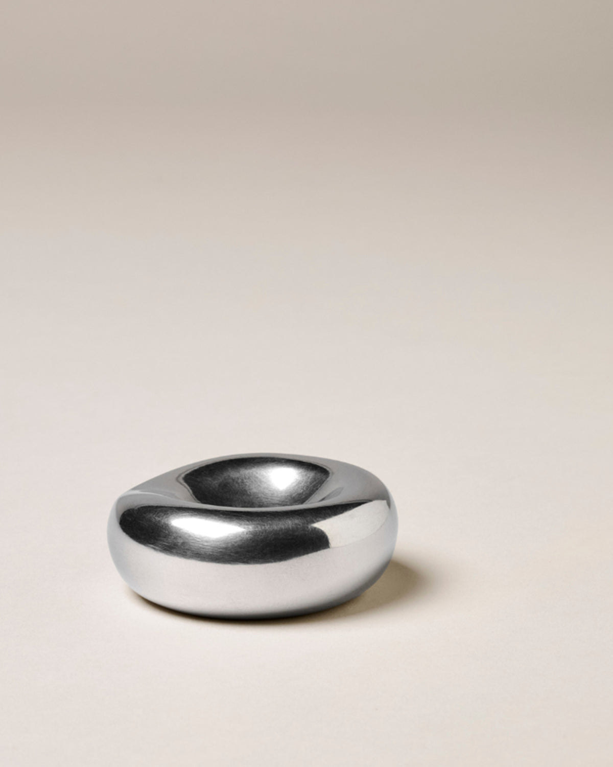 A minimalist, organic-shaped chrome incense holder with a smooth, polished finish, set against a solid warm beige background.
