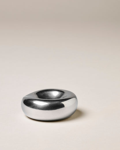 A minimalist, organic-shaped chrome incense holder with a smooth, polished finish, set against a solid warm beige background.