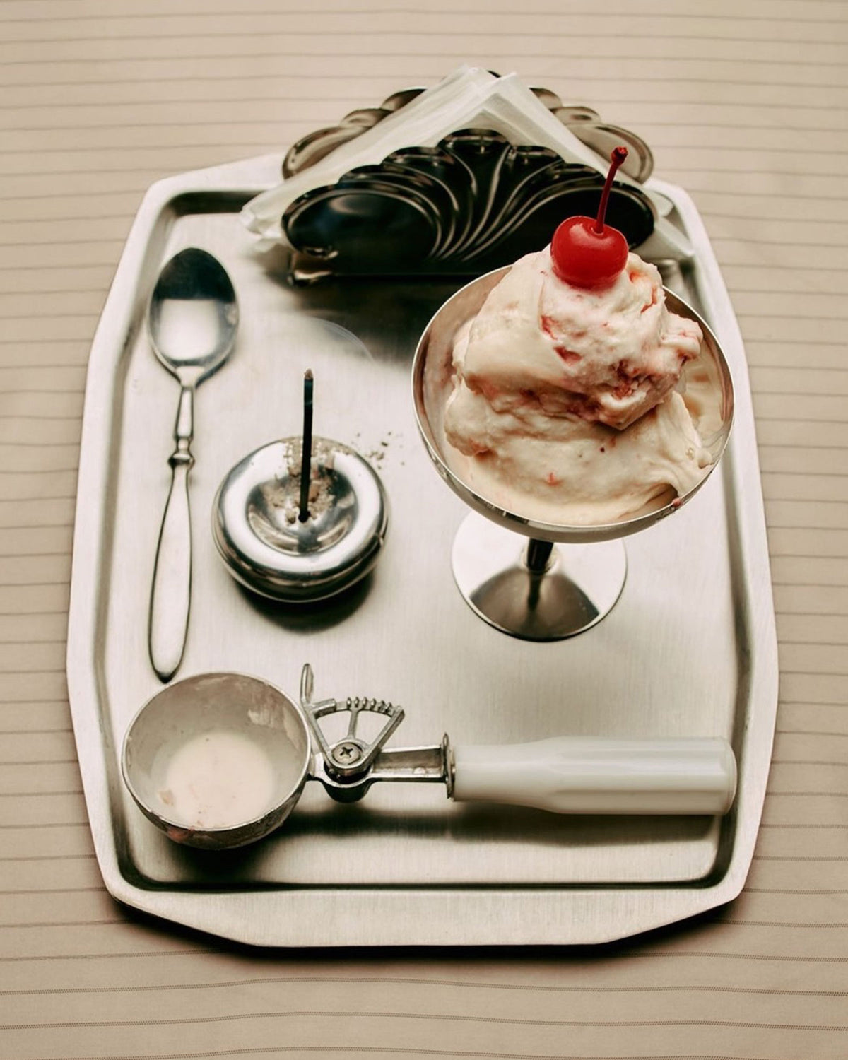 A dessert presentation on a metal tray, featuring a scoop of vanilla ice cream topped with a red cherry in a stemmed metal martini-shaped vessel, alongside a metal ice cream scoop, metal spoon, metal serviette holder and bowl-shaped metal incense holder holding a vertical incense stick which is lit and smouldering, ash remnants are scattered in and around the incense holder. 