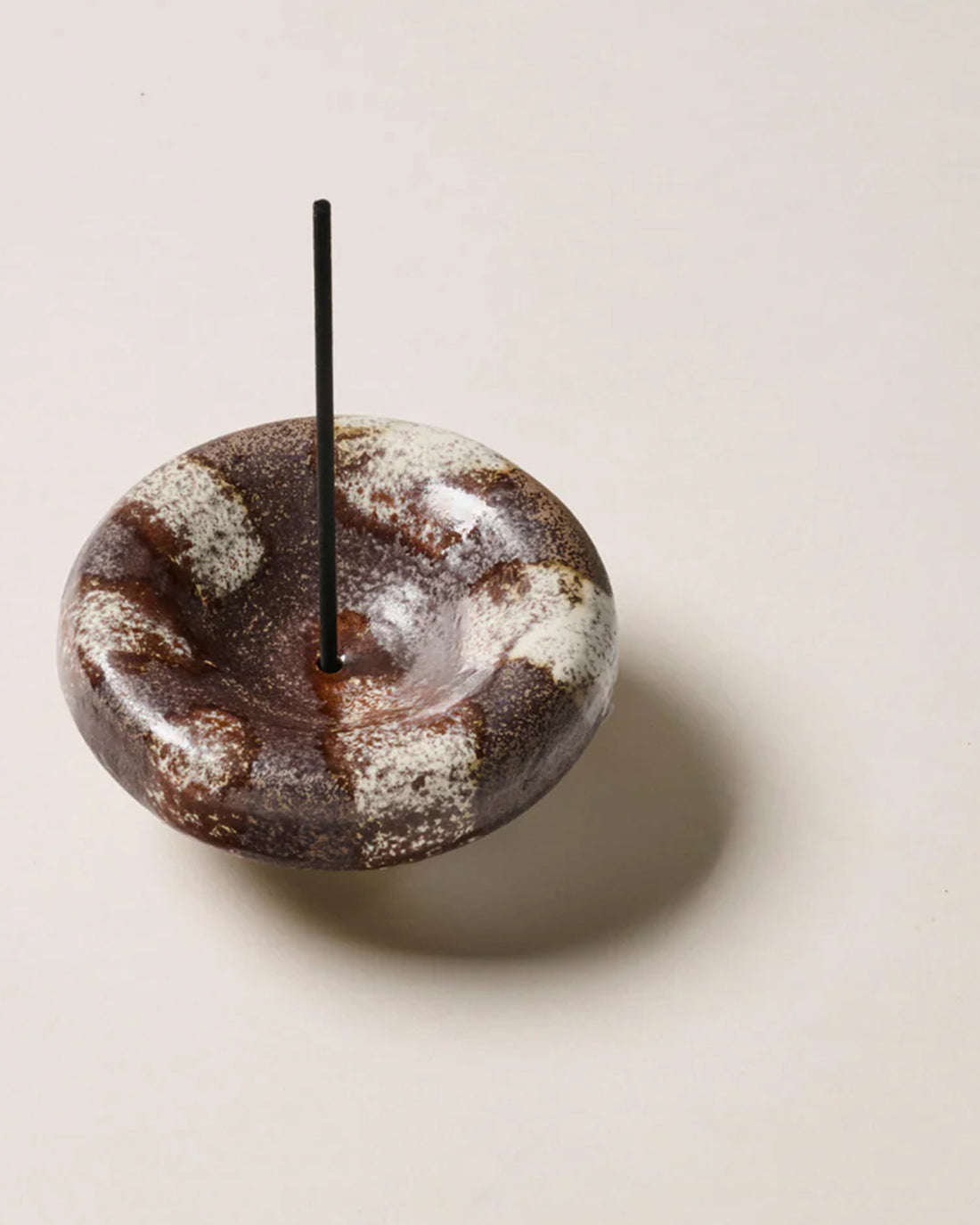 A round, bowl-shaped ceramic incense holder, painted brown with organic off-white stripes, one black incense stick sits vertically in the holder. The incense holder sits alone on an off-white background.