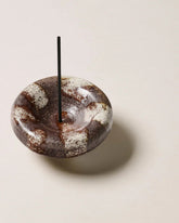 A round, bowl-shaped ceramic incense holder, painted brown with organic off-white stripes, one black incense stick sits vertically in the holder. The incense holder sits alone on an off-white background.
