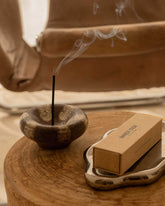 A styled sidetable features a brown and beige ceramic incense holder holding a burning incense stick next to an ornate silver plate holding a box of incense.