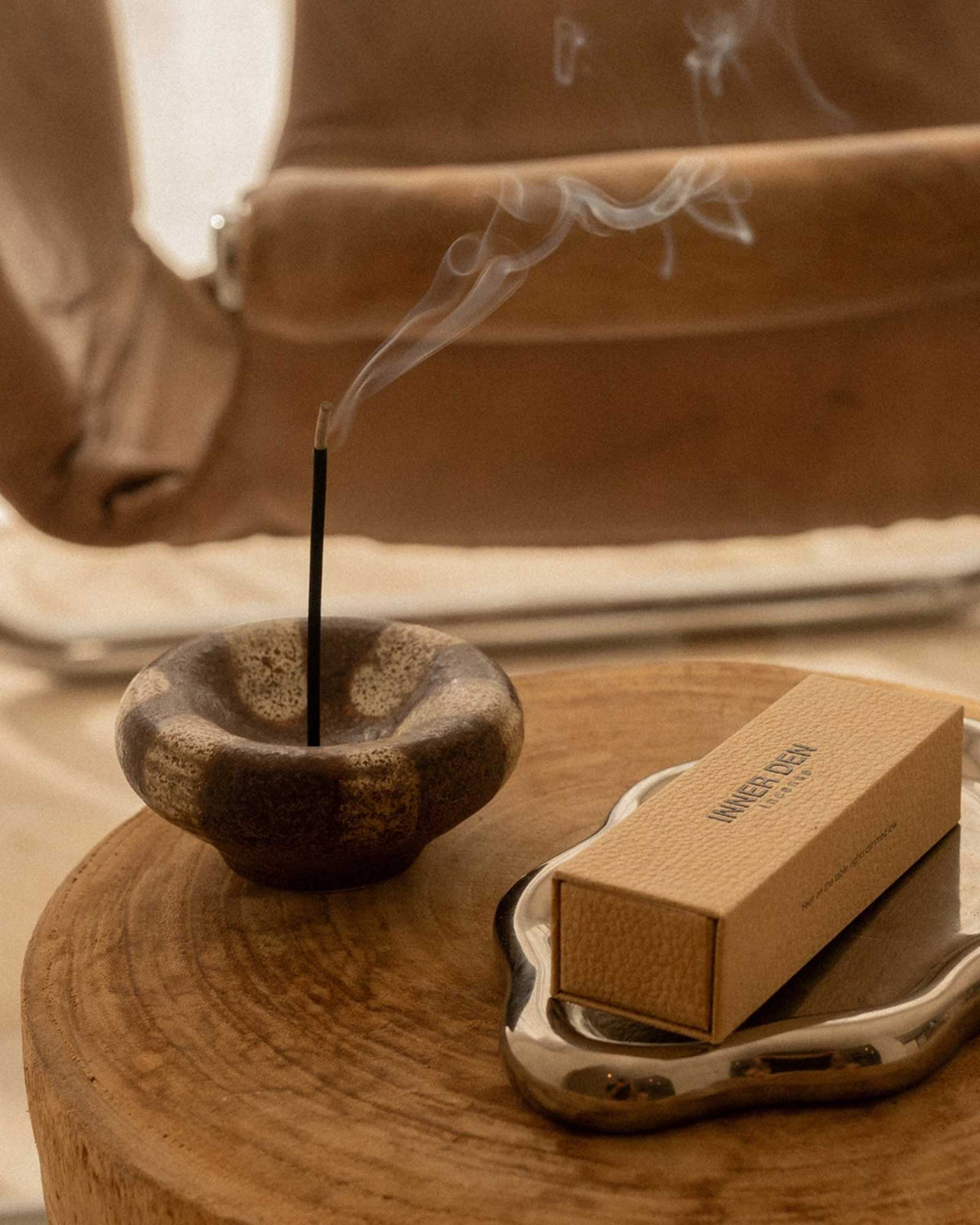 A styled sidetable features a brown and beige ceramic incense holder holding a burning incense stick next to an ornate silver plate holding a box of incense.