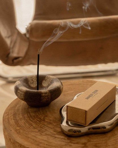 A styled sidetable features a brown and beige ceramic incense holder holding a burning incense stick next to an ornate silver plate holding a box of incense.