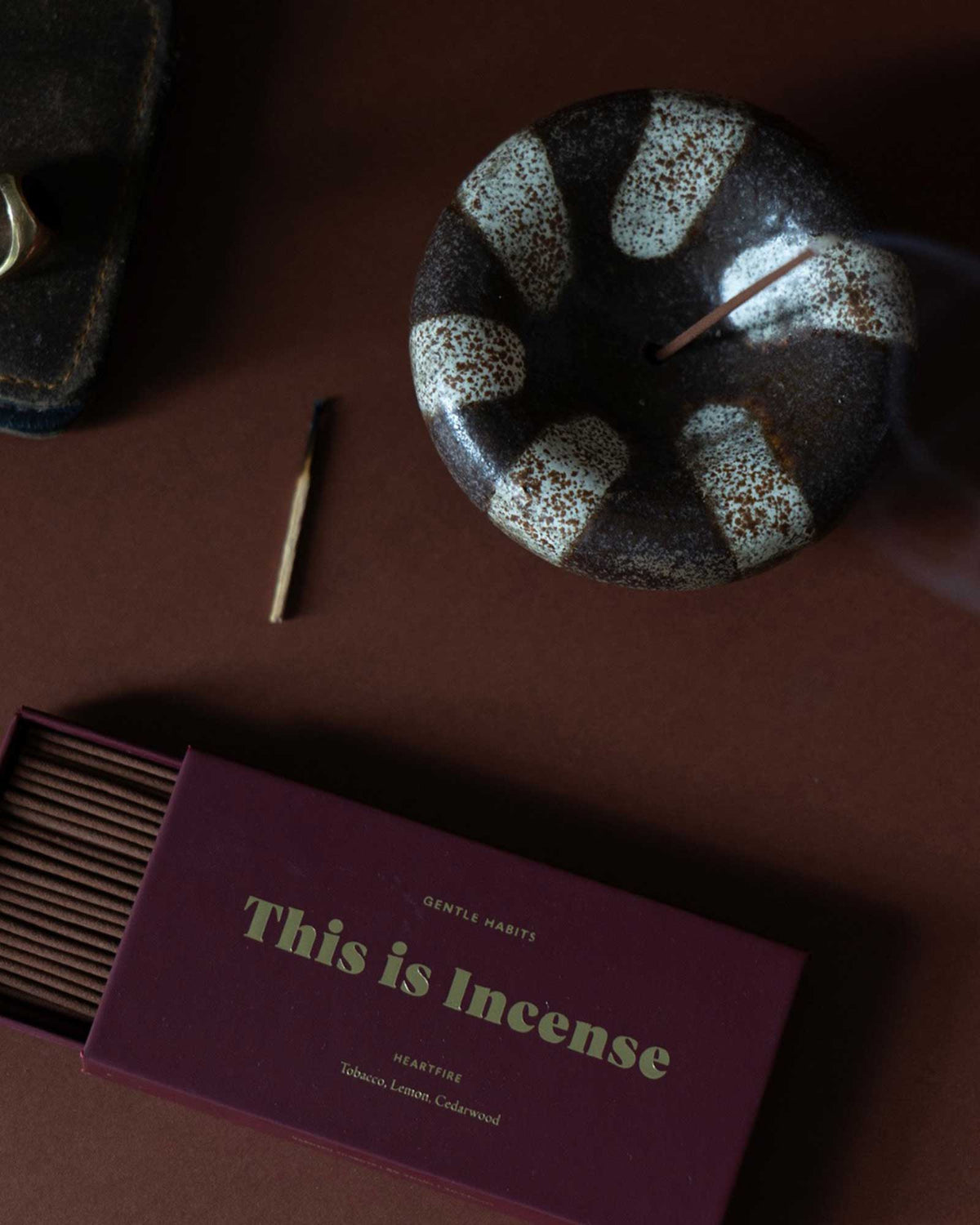 A close-up, aerial view of scattered items on a dark table including a round, bowl-shaped ceramic incense holder holding a lit and smouldering incense stick, a burnt matchstick and an open box of incense sticks. 