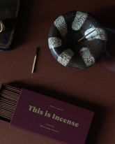 A close-up, aerial view of scattered items on a dark table including a round, bowl-shaped ceramic incense holder holding a lit and smouldering incense stick, a burnt matchstick and an open box of incense sticks. 