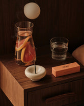 A styled product scene on a dark wood nightstand featuring a white ceramic incense holder with a burning incense stick, a peach-coloured incense box, a glass of water, and a glass carafe with grapefruit slices, set against a dark wood background.