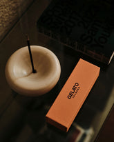 A white ceramic bowl-shaped incense holder containing a single incense stick sits on a dark reflective tabletop alongside a box of incense.