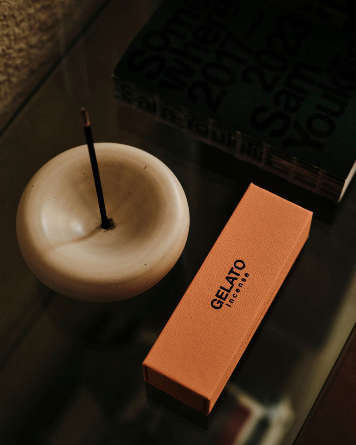 A white ceramic bowl-shaped incense holder containing a single incense stick sits on a dark reflective tabletop alongside a box of incense.