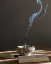 A ceramic incense holder vertically holds a burning stick of incense, a stream of smoke flows from the tip of the incense into the air. 