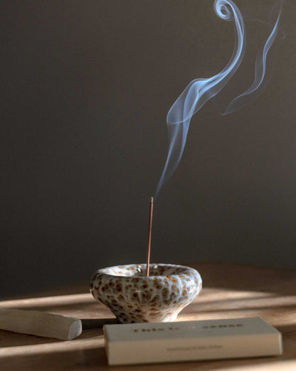 A ceramic incense holder vertically holds a burning stick of incense, a stream of smoke flows from the tip of the incense into the air. 