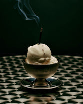 A metal dessert bowl filled with 2 scoops of white ice cream with a burning incense stick poking out from the top ice cream scoop. The incense tip is lit and a line of smoke is drifting from the stick. The dessert bowl sits alone on an old fashioned cafe table against a dark green wall.
