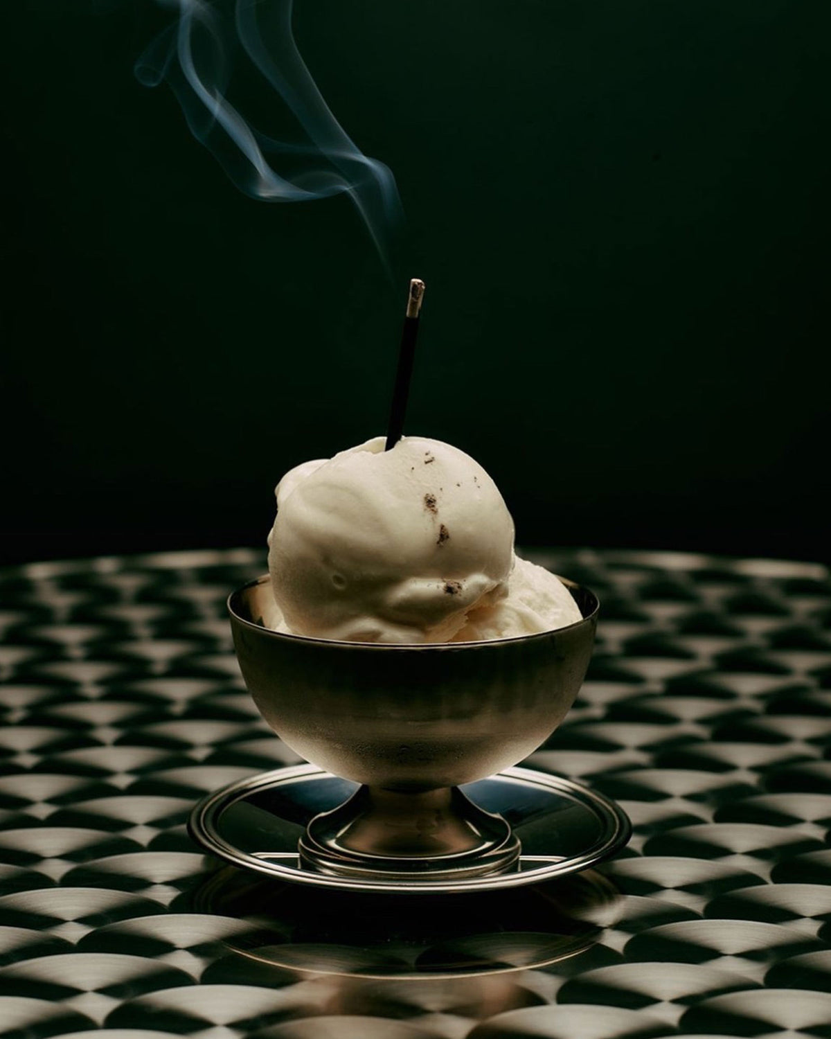 A metal dessert bowl filled with 2 scoops of white ice cream with a burning incense stick poking out from the top ice cream scoop. The incense tip is lit and a line of smoke is drifting from the stick. The dessert bowl sits alone on an old fashioned cafe table against a dark green wall.