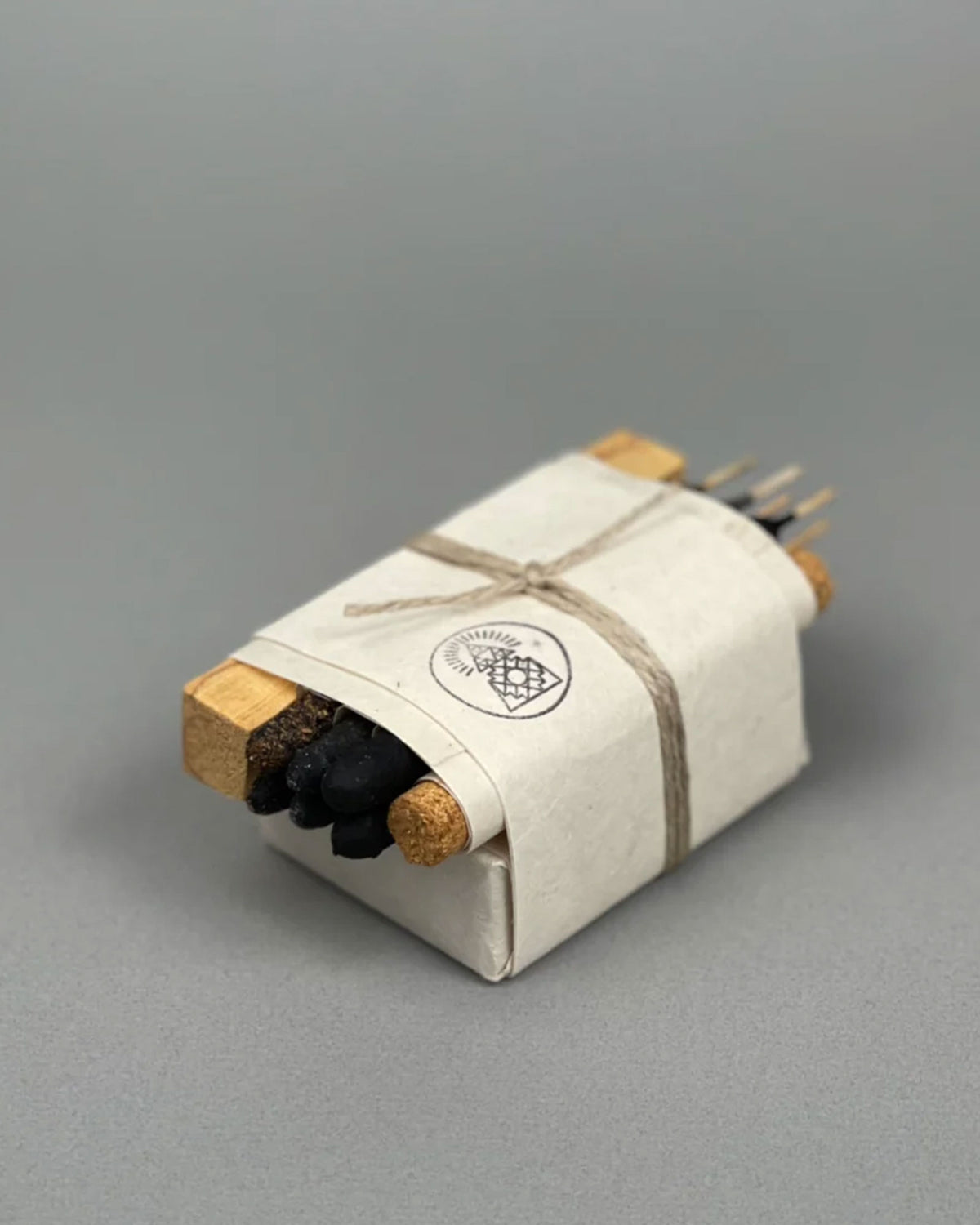 A wrapped bundle of incense sticks tied with twine, showing light wood ends, dark charcoal sticks, and the Incausa logo on the paper wrap, photographed on a neutral grey, seamless background.