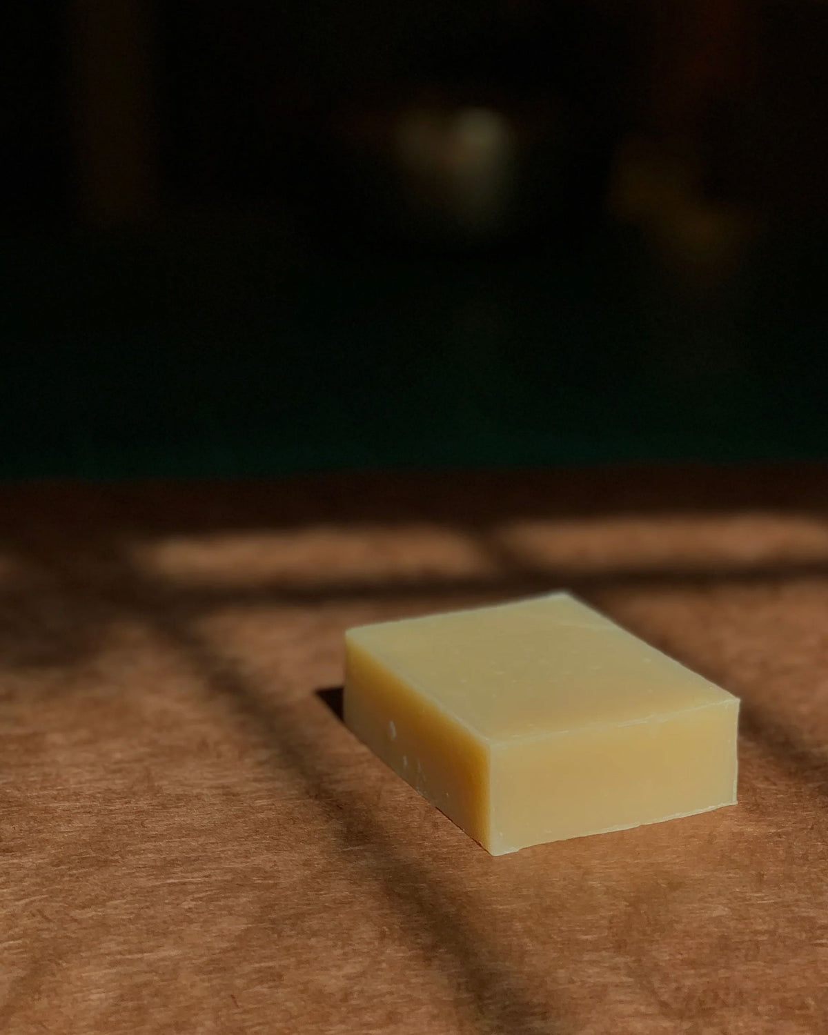 A rectangular bar of pale yellow soap bar resting on a textured brown surface with soft shadows cast across it, against a dark blurred background.