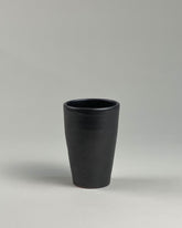 A tall black ceramic drinking tumbler on a seamless, light grey background. 