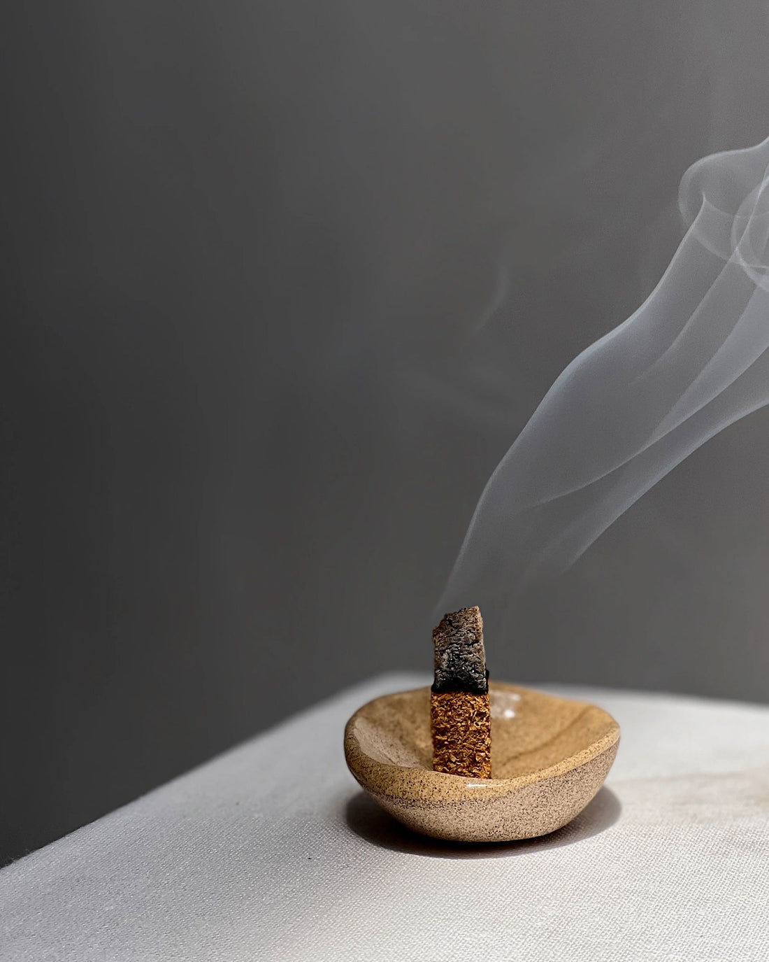 A small brown organic shaped bowl holding a smouldering stick of compressed wood which is smouldering with a long stream of smoke from the tip. Set on a white table surface with a grey background. 