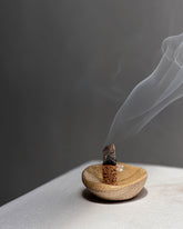 A small brown organic shaped bowl holding a smouldering stick of compressed wood which is smouldering with a long stream of smoke from the tip. Set on a white table surface with a grey background. 