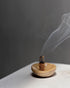 A small brown organic shaped bowl holding a smouldering stick of compressed wood which is smouldering with a long stream of smoke from the tip. Set on a white table surface with a grey background. 