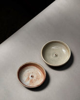 Two round, bowl-like, ceramic incense holders sit side-by-side on a light grey table surface. Both incense holders are earthy tones, the one on the left is a mx of brown, white and red earth, the holder on the right is a mix of light sage and stone colours. 