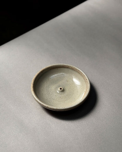 A natural-coloured, round, bowl-like, ceramic incense holder sits on a light grey table surface.