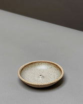A natural-coloured, round, bowl-like, ceramic incense holder sits on a light grey table surface.