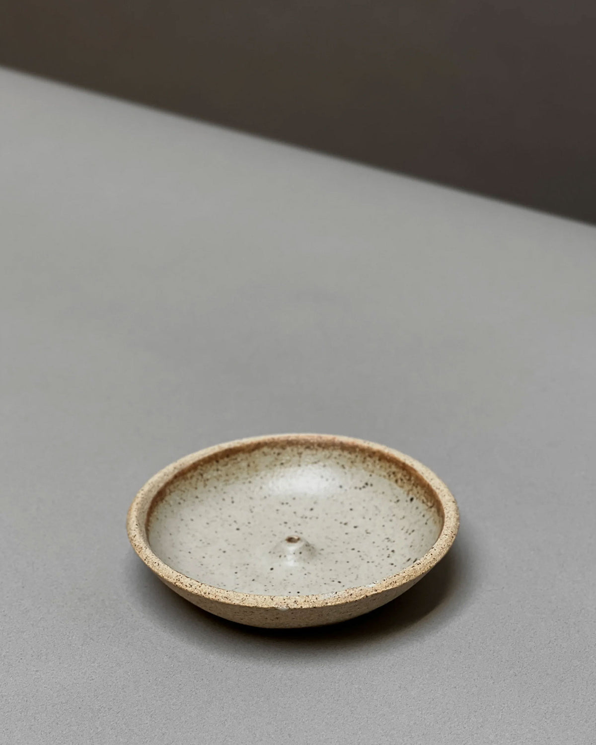 A natural-coloured, round, bowl-like, ceramic incense holder sits on a light grey table surface.