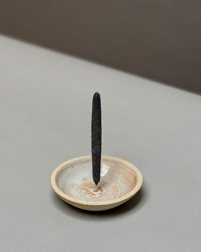 A natural-coloured, round, bowl-like, ceramic incense holder with a large charcoal incense stick held vertically from the centre of the holder sits on a light grey table surface.