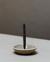 A natural-coloured, round, bowl-like, ceramic incense holder with a large charcoal incense stick held vertically from the centre of the holder sits on a light grey table surface.
