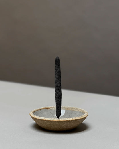 A natural-coloured, round, bowl-like, ceramic incense holder with a large charcoal incense stick held vertically from the centre of the holder sits on a light grey table surface.